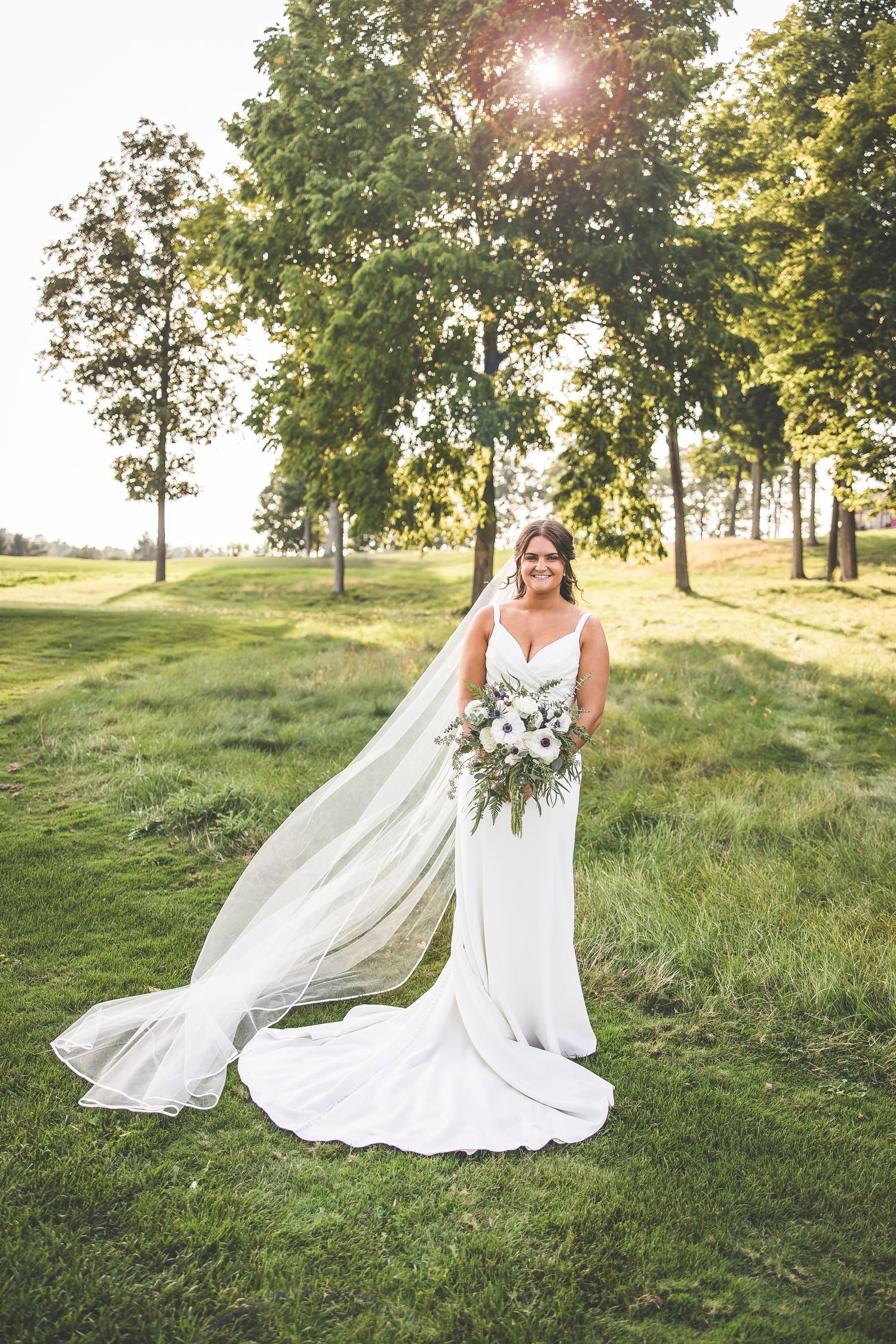 Bride in flowing white dress poses in sunlit meadow with flowing veil and bouquet.