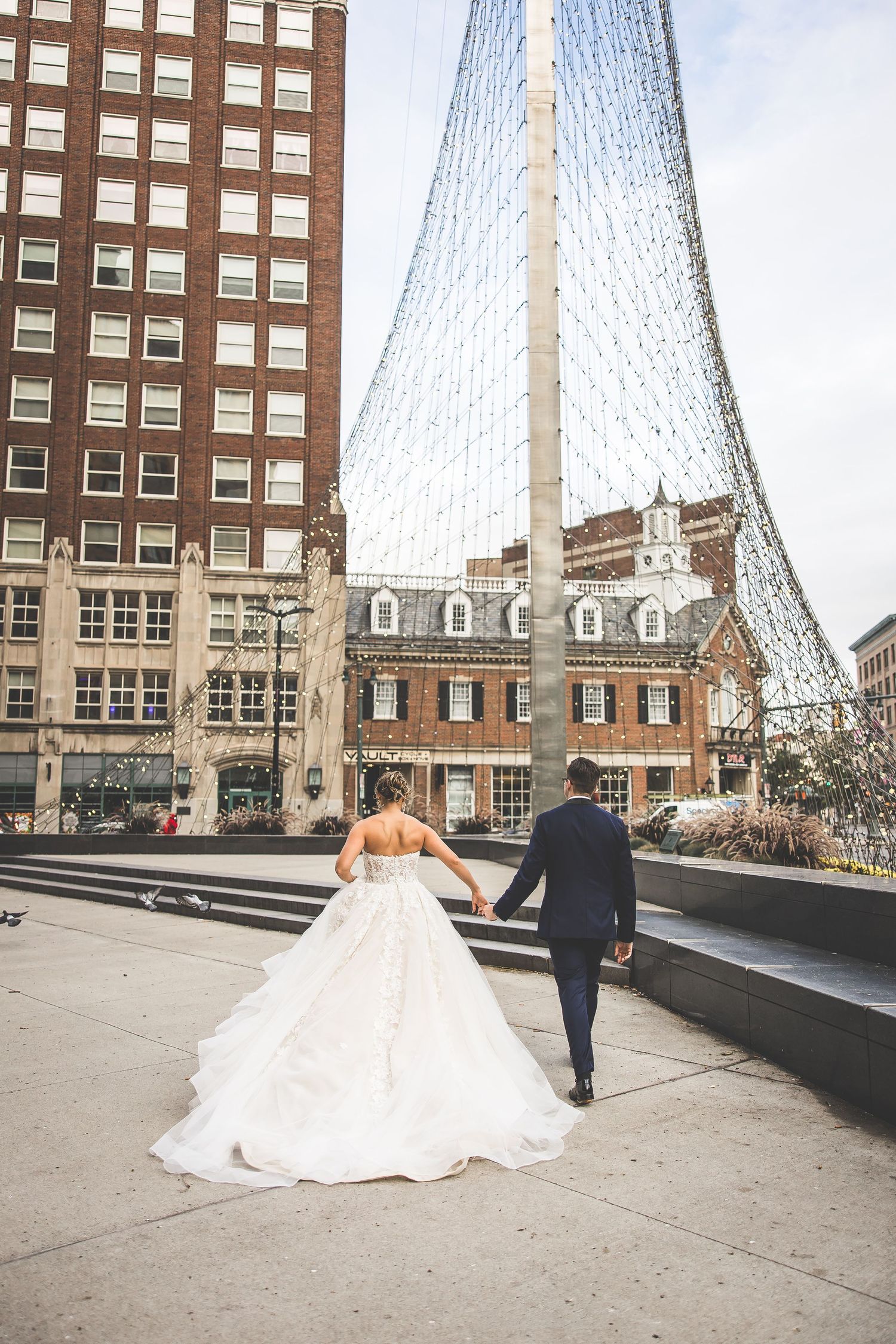 Couple in wedding attire walks hand in hand near modern glass building structure.