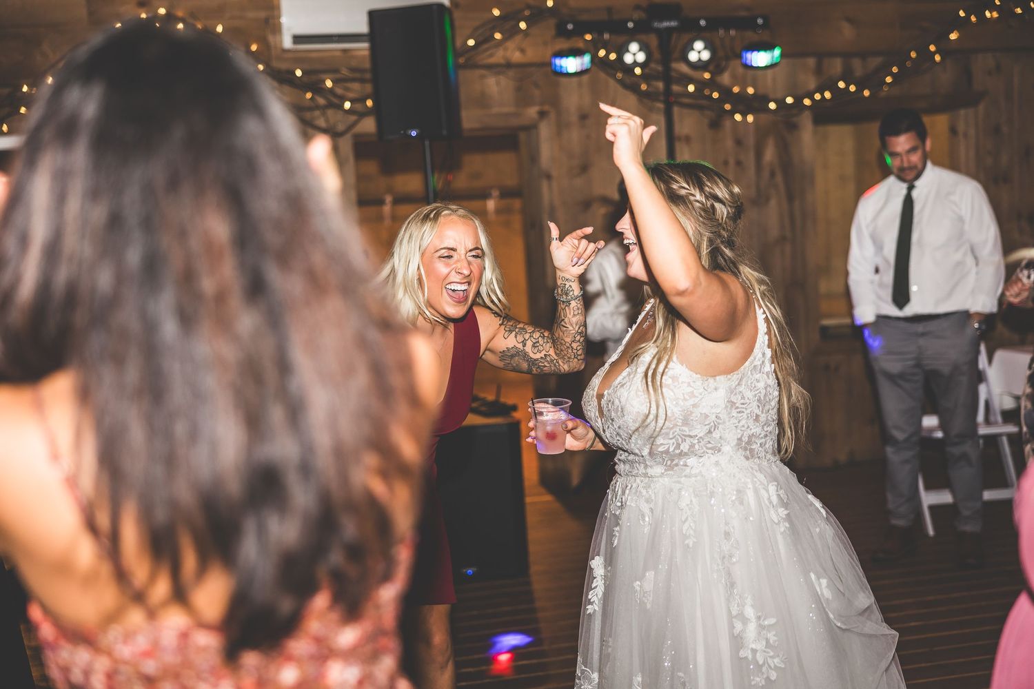 Wedding celebration in full swing with guests dancing under twinkling lights in a barn venue.