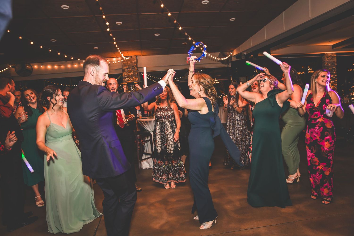 Wedding guests dance enthusiastically on a dimly lit dance floor decorated with string lights.