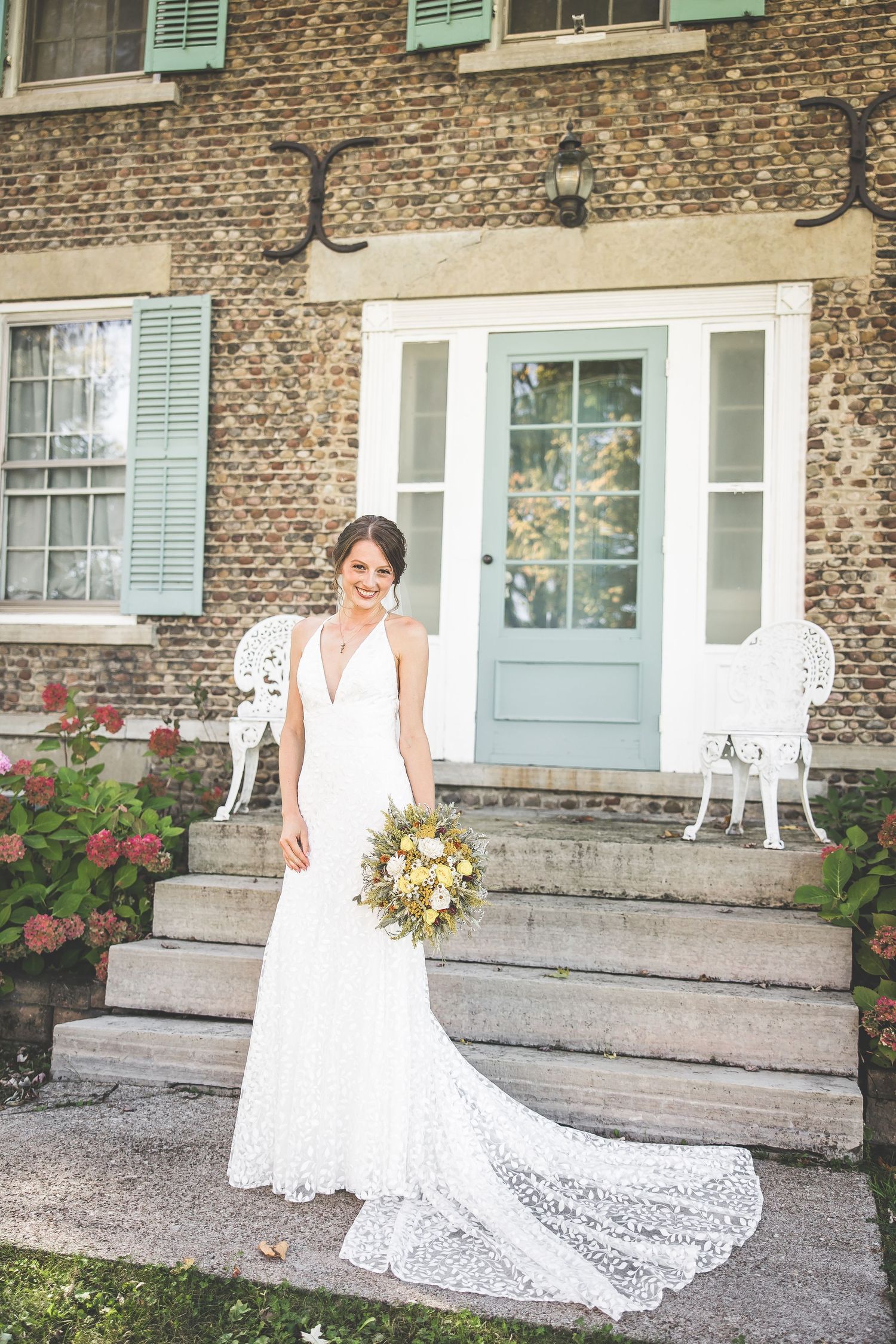 A bride in a flowing lace wedding gown poses on stone steps of a rustic building with white iron chairs.