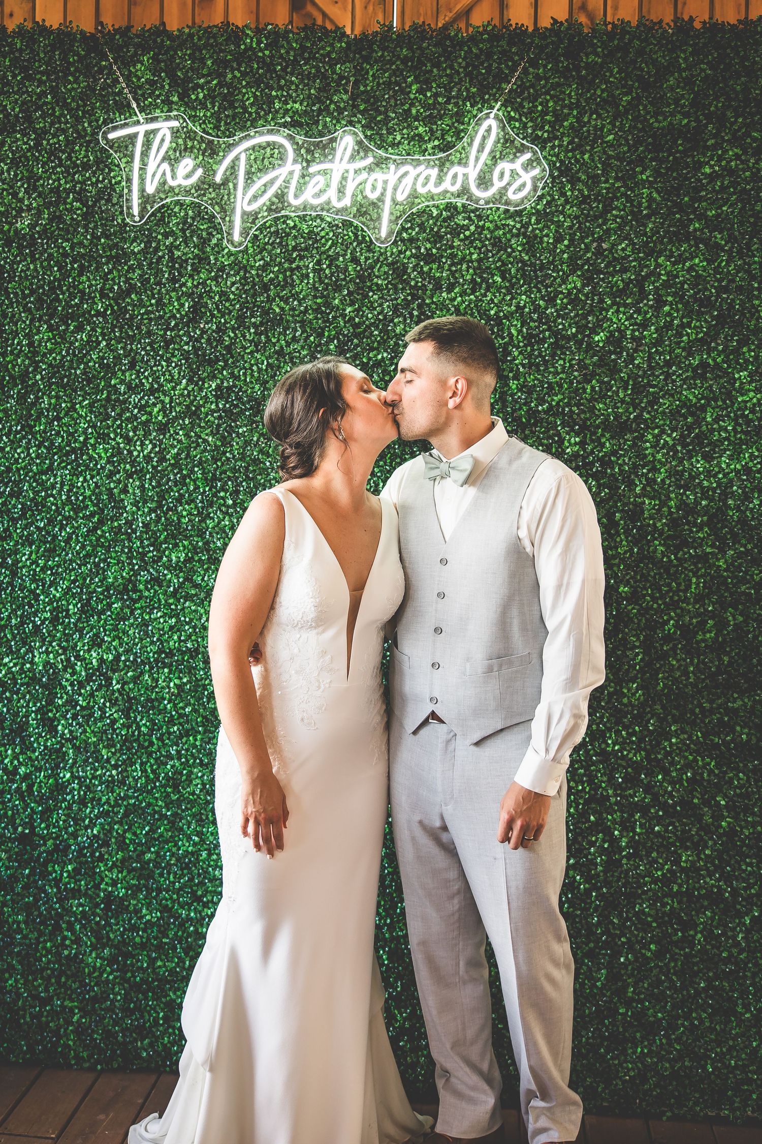 Romantic couple poses against a green hedge wall with illuminated script signage.