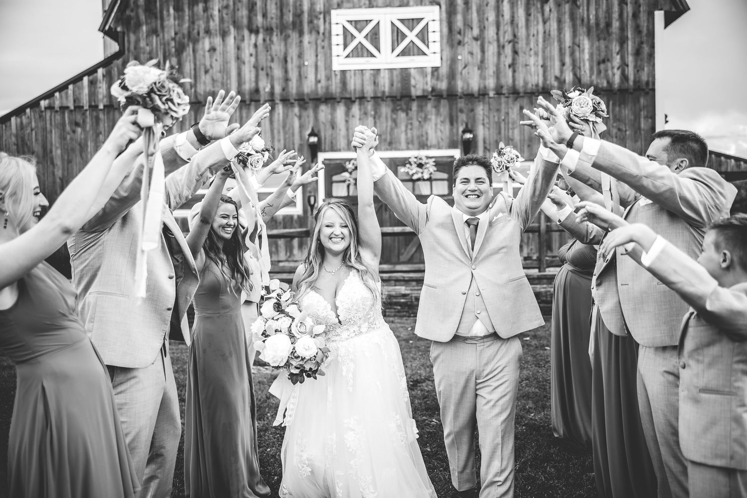 Wedding party celebrates with energetic group poses outside rustic barn venue.