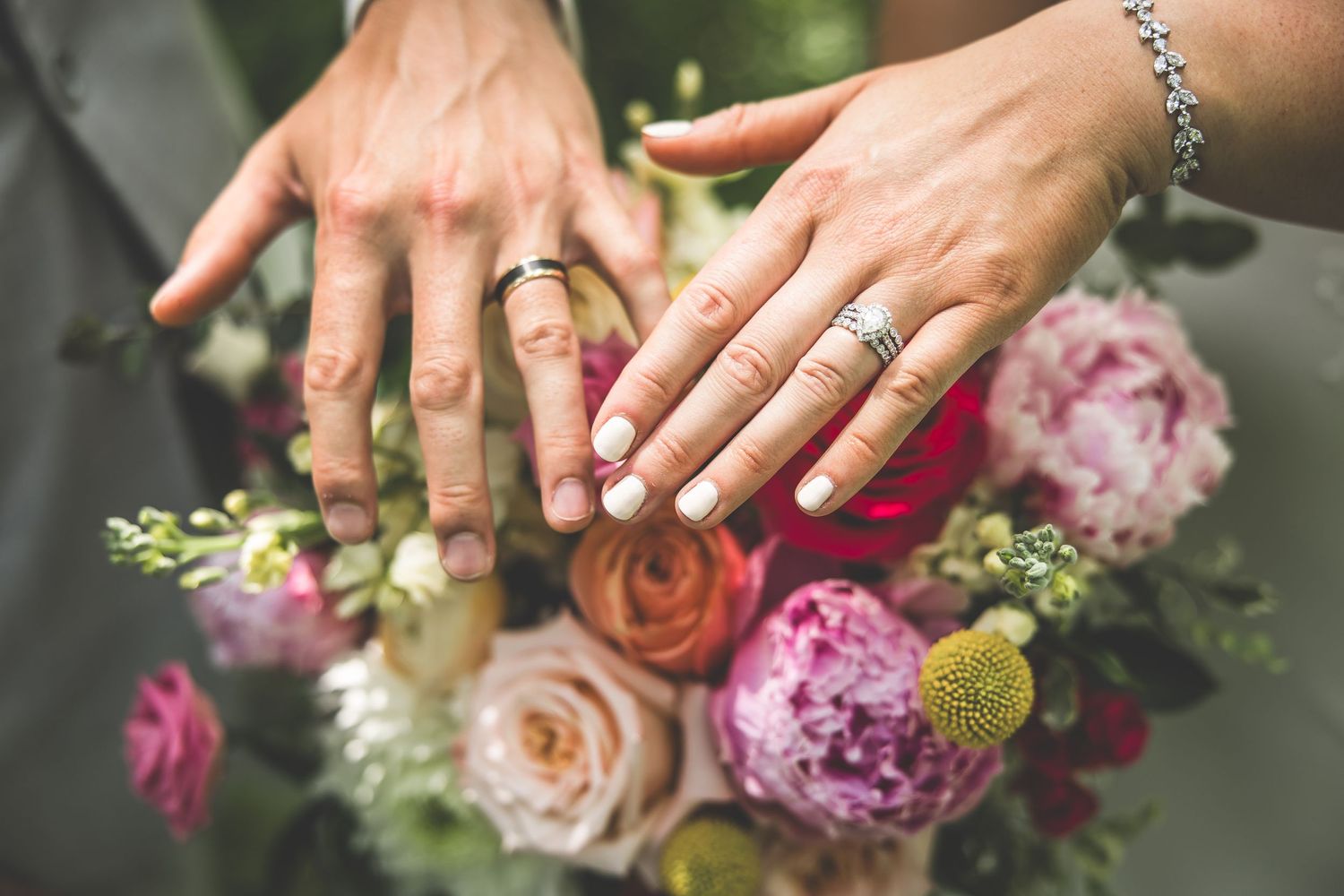 Close-up of wedding rings and hands against a vibrant bouquet of pink and peach roses.