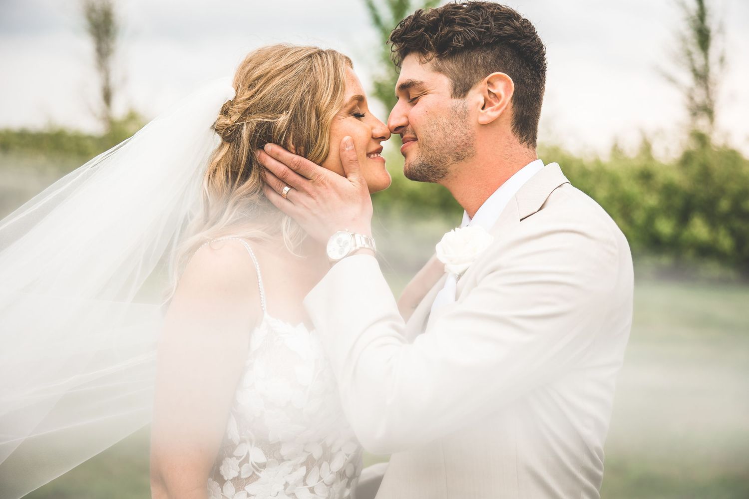 Intimate wedding couple portrait with flowing veil against natural outdoor backdrop.