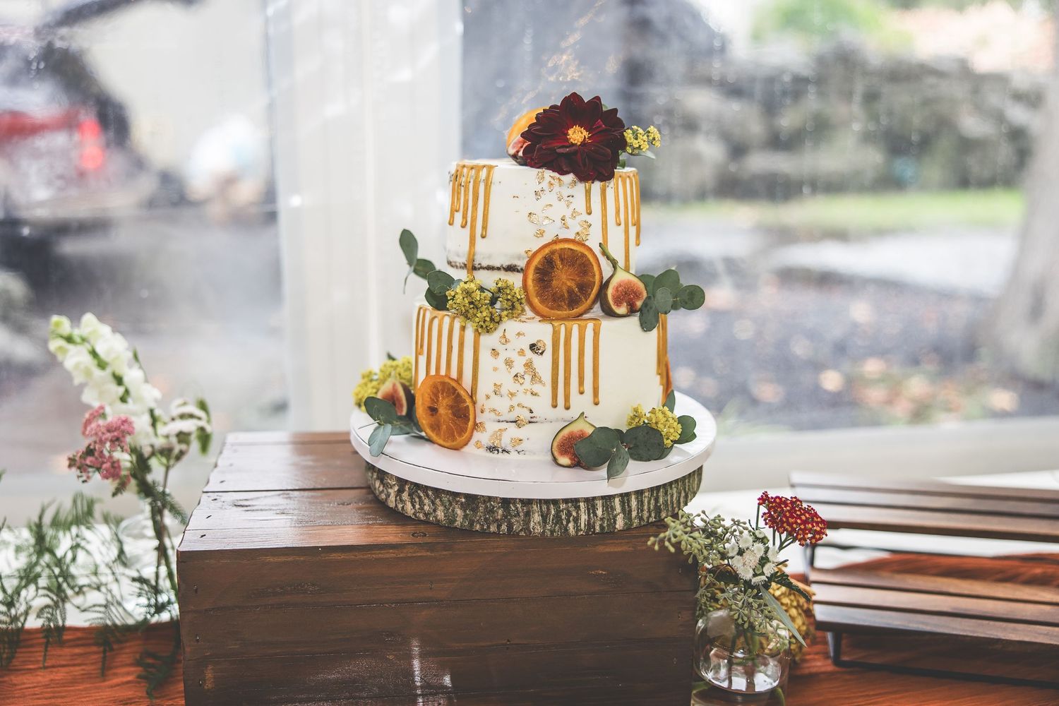 Two-tiered white wedding cake decorated with fall-colored flowers and greenery on a wood slice stand.