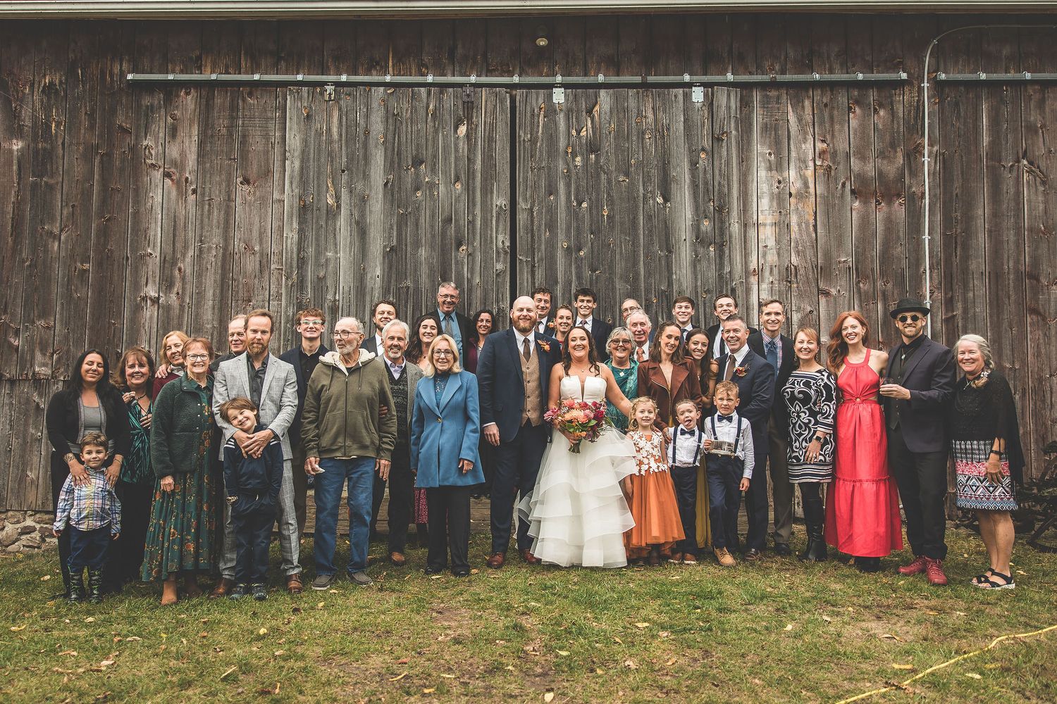 Large group wedding photo of guests lined up against rustic barn exterior.