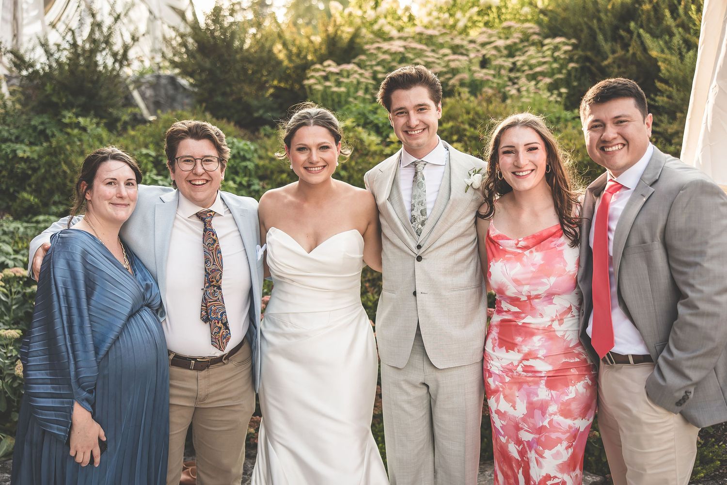Wedding party poses together outdoors in formal attire with light gray suits and coral dresses.