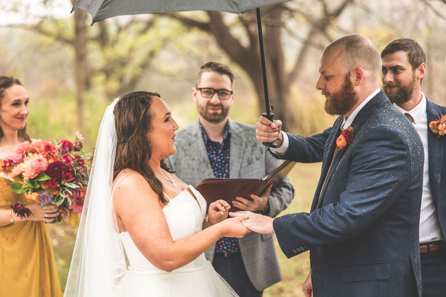Couple exchanges rings during outdoor autumn wedding ceremony with umbrella overhead.