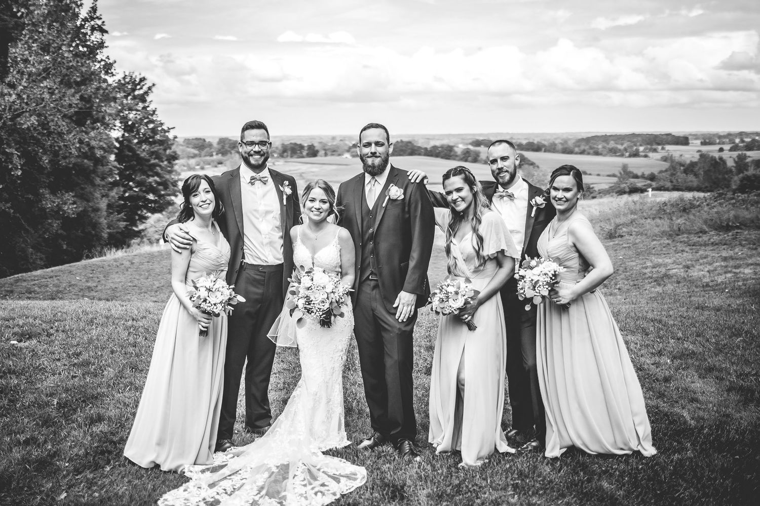 Wedding group photo with bridesmaids in light blue and groomsmen in navy suits on rolling hills.