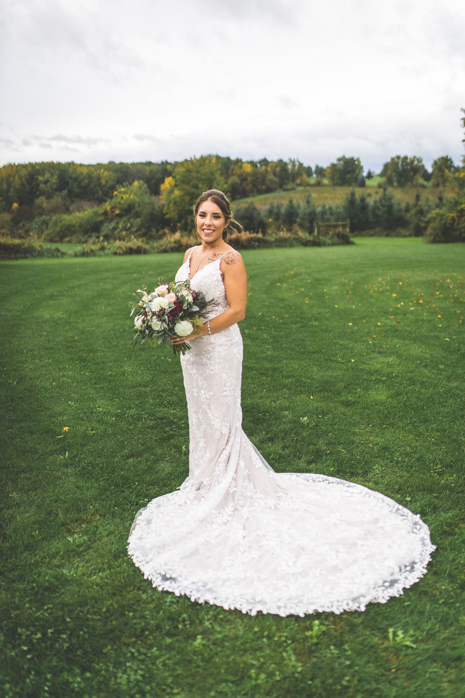 Bride in lace wedding gown with long train poses in green outdoor field setting.