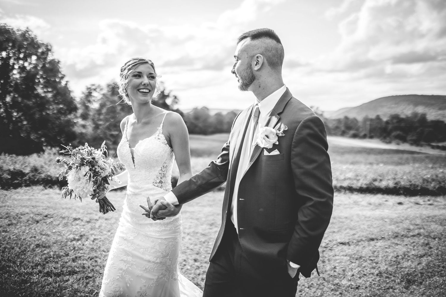 A couple embraces while standing in a scenic outdoor field on their wedding day.