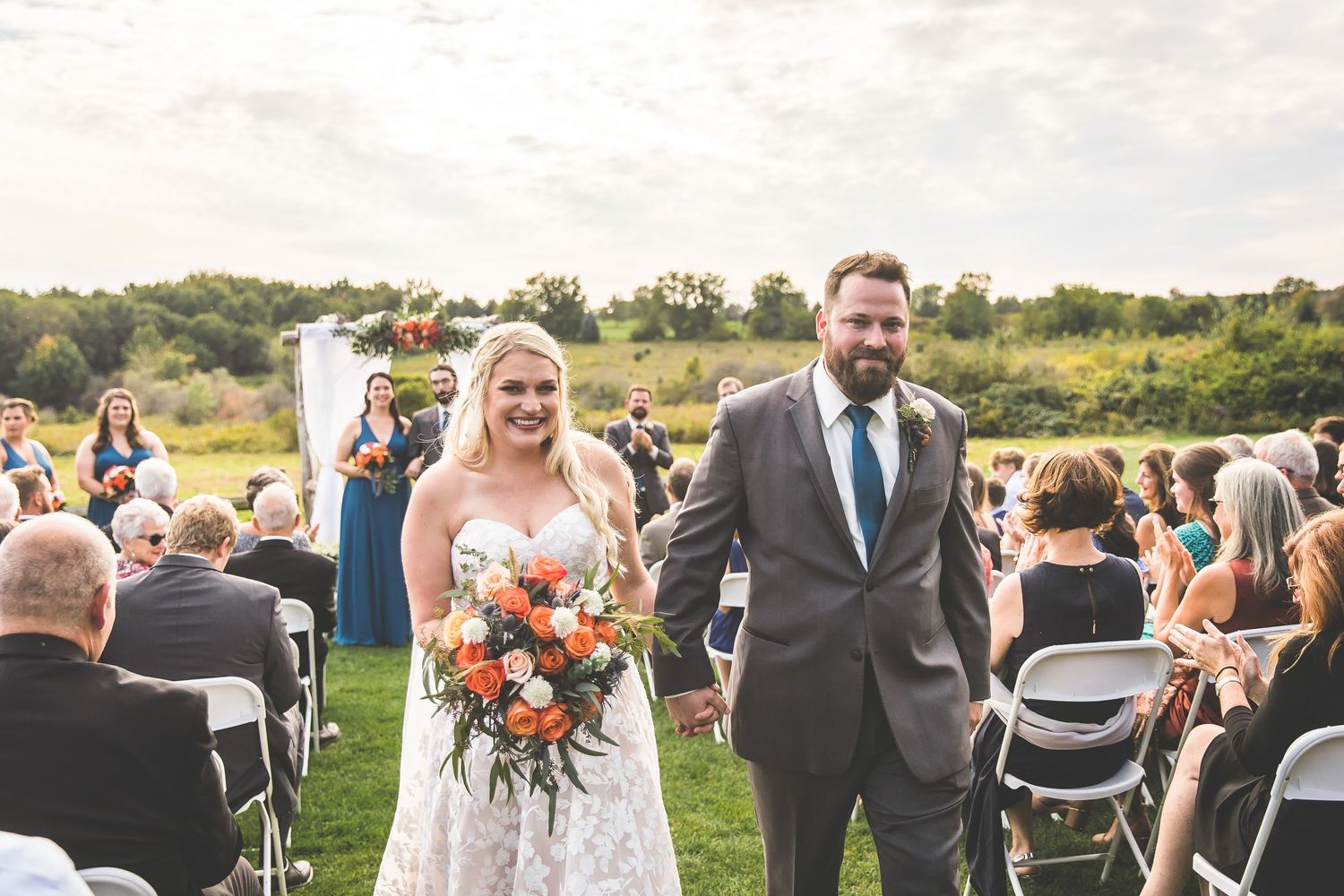 Wedding celebration outdoors with guests gathered on grassy field under blue sky.