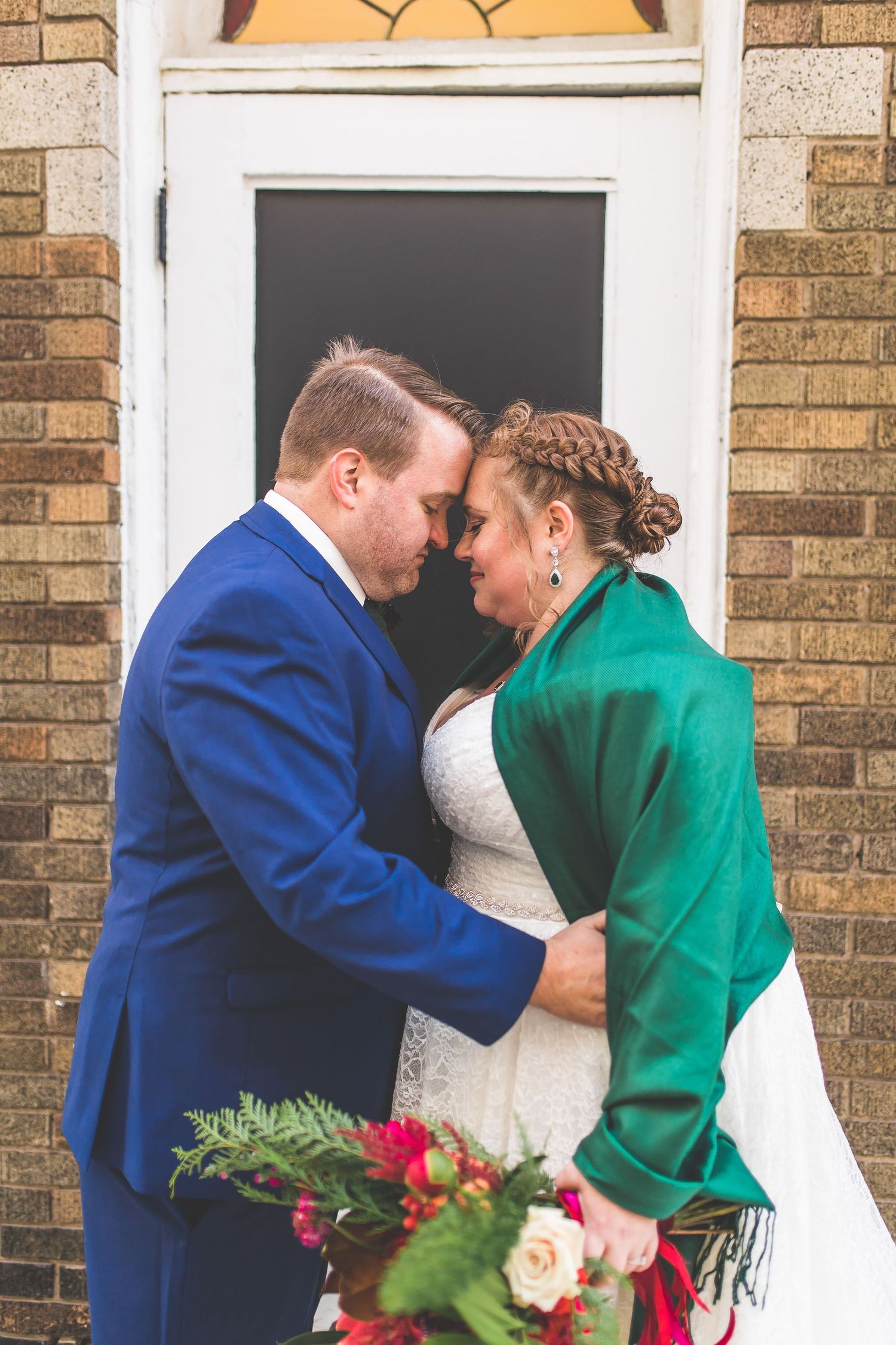 Intimate wedding portrait of couple by brick doorway with red and white bouquet.