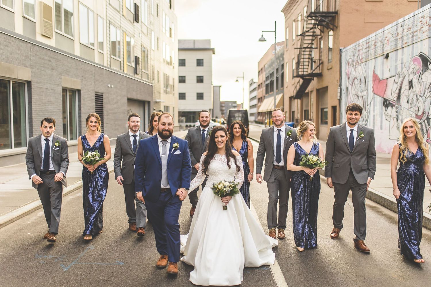 Wedding party walks down urban street with bridesmaids in navy and groomsmen in suits.