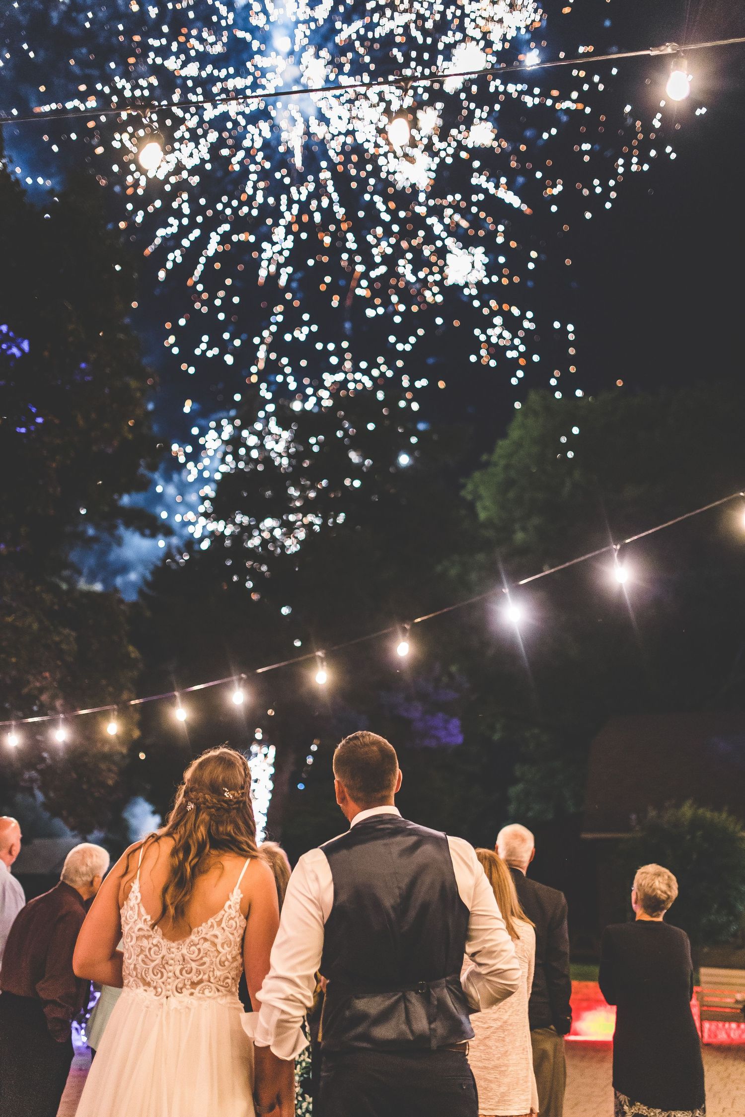 Fireworks light up the night sky over a wedding celebration with string lights below.