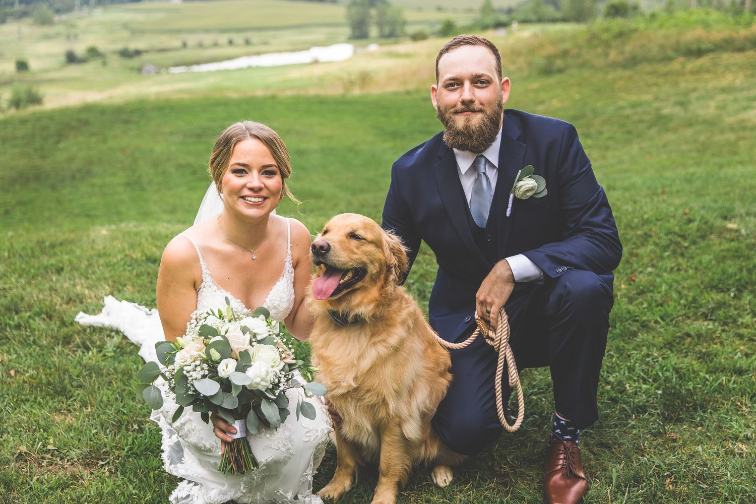 Bride and groom kneel beside their golden retriever for casual wedding portrait outdoors.