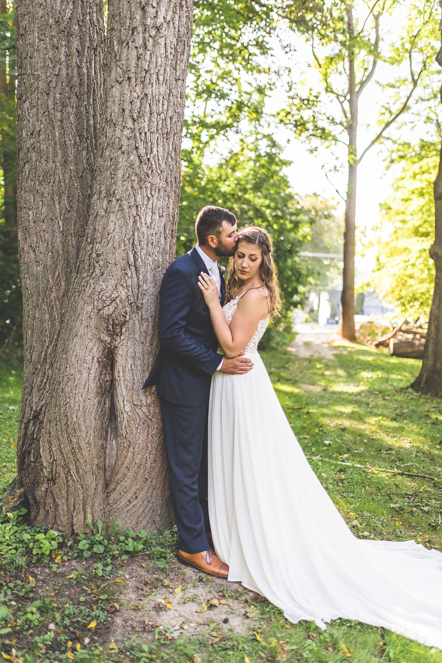 A romantic wedding couple poses against a large tree trunk in a sunlit garden setting.
