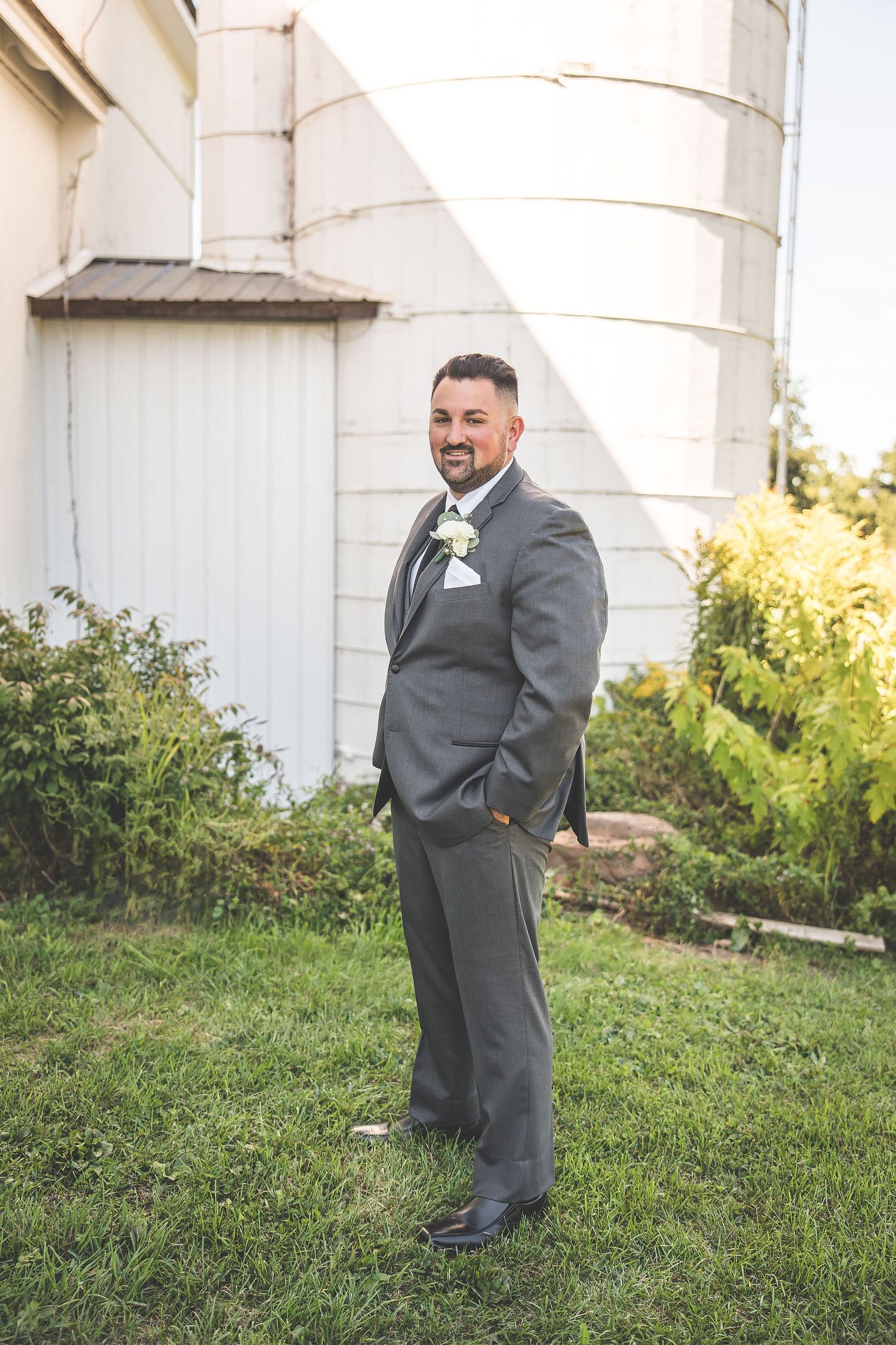 Groom in grey suit poses by white silo on grassy lawn during wedding photoshoot.