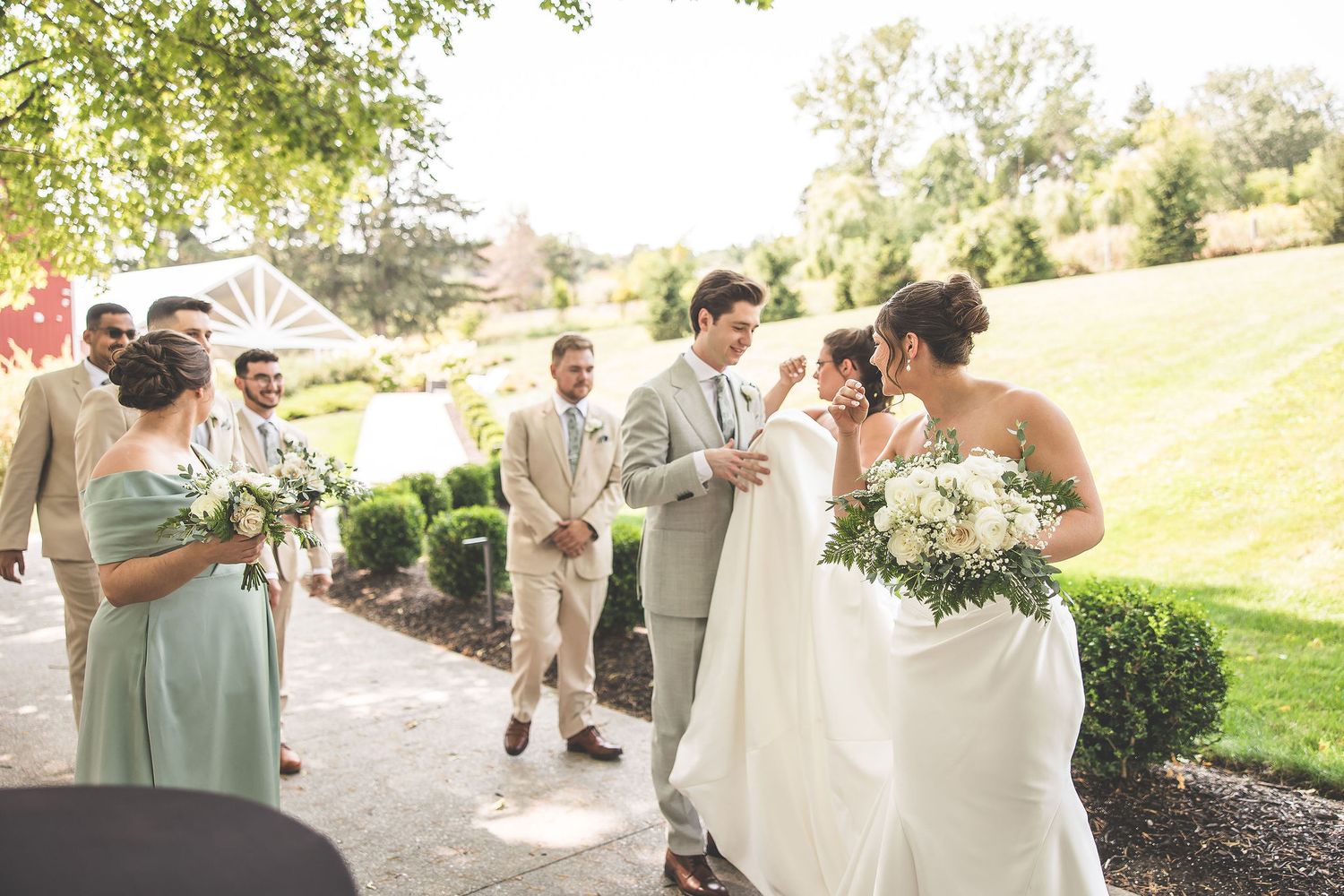Wedding party poses for photos on a sunny day wearing light colored attire and holding bouquets.