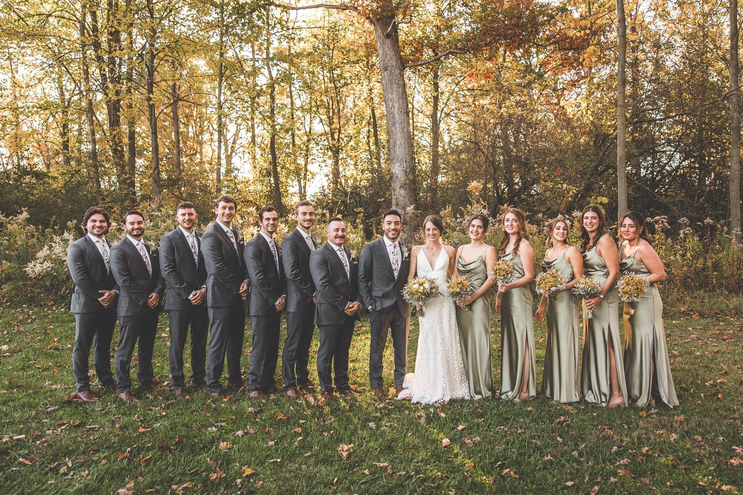 Wedding party poses in autumn forest with golden sunlight filtering through trees.