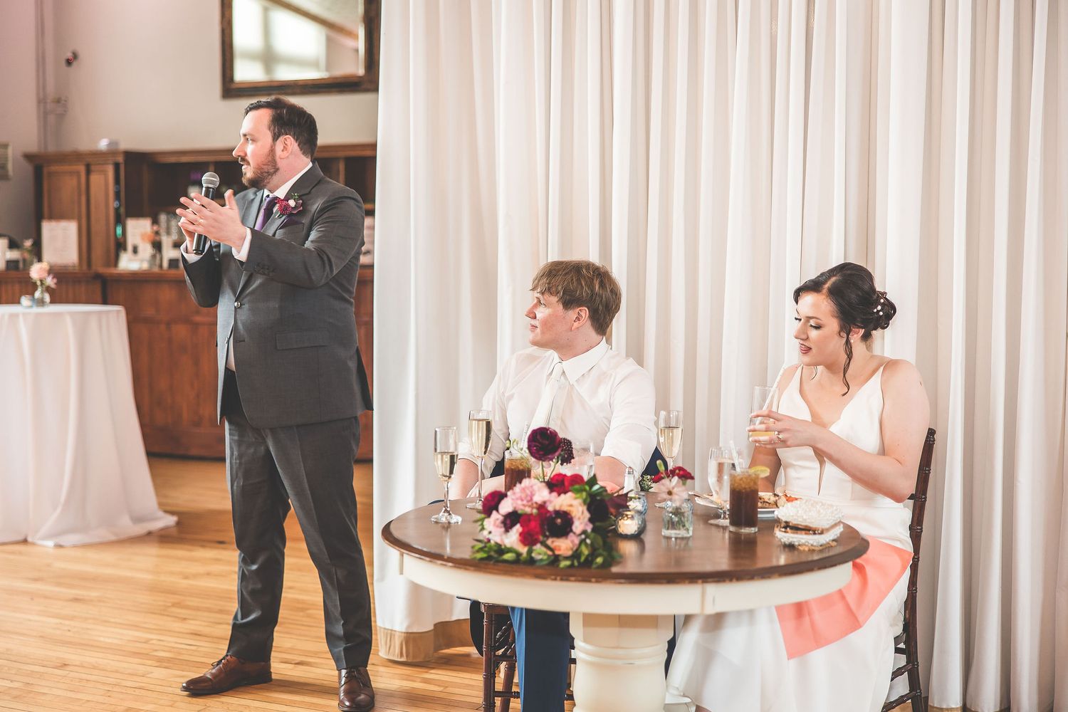Wedding guests mingle during reception in a bright room with flowing white curtains.