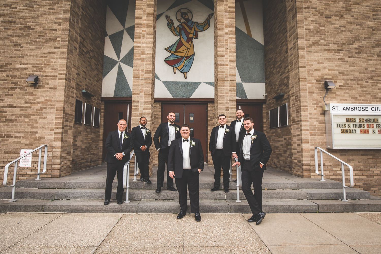 Wedding party in formal attire poses on the steps of a brick church with decorative artwork.