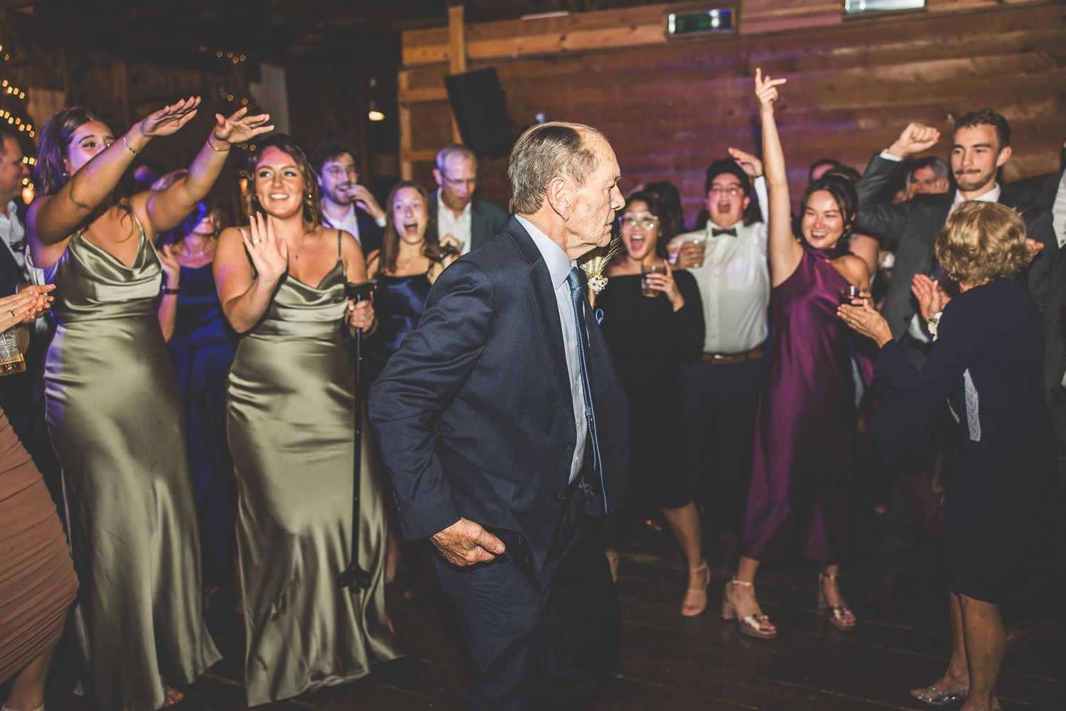 Wedding party members in formal attire dancing in line at rustic barn venue.