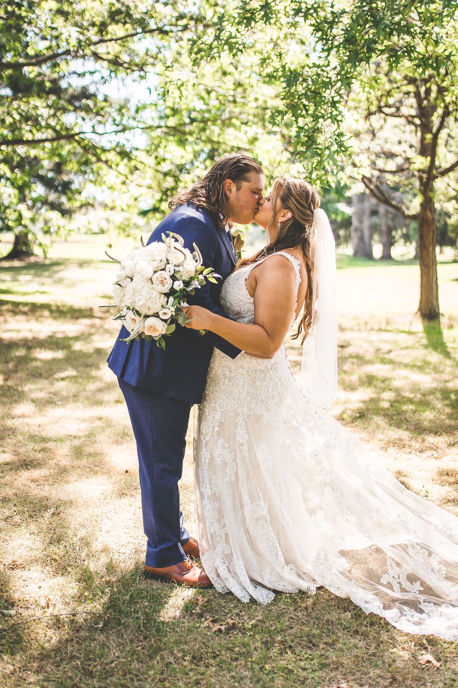 Bride in lace dress and groom in navy suit share a romantic moment under trees.