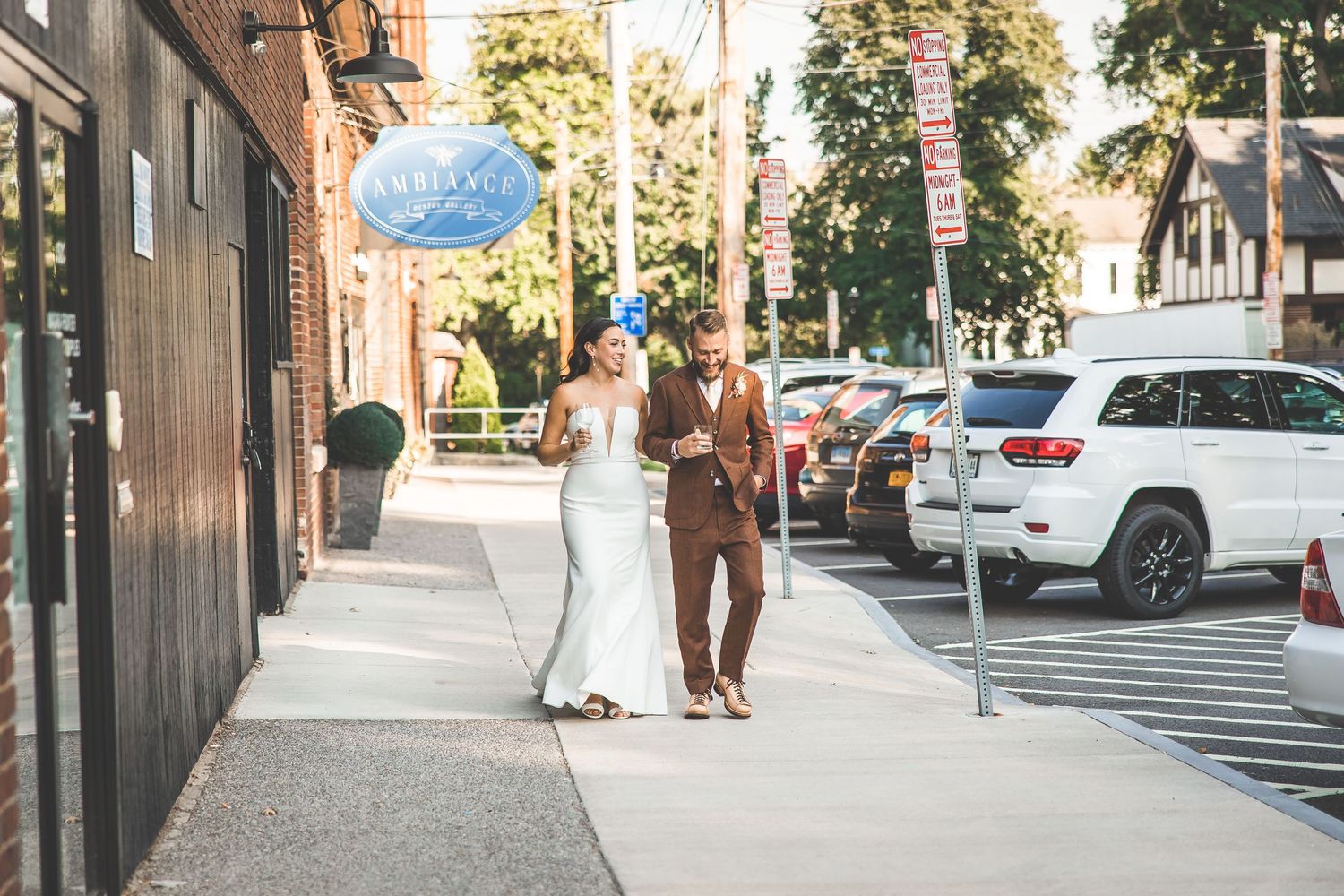 A couple walks down a city sidewalk on a sunny day with parked cars and trees in the background.