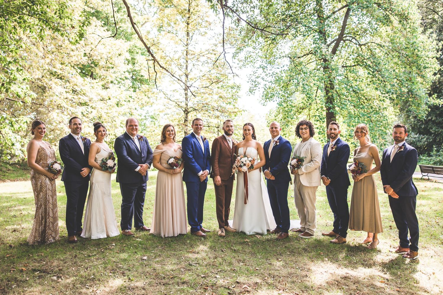 Wedding party members arranged in a long line under green tree canopy.