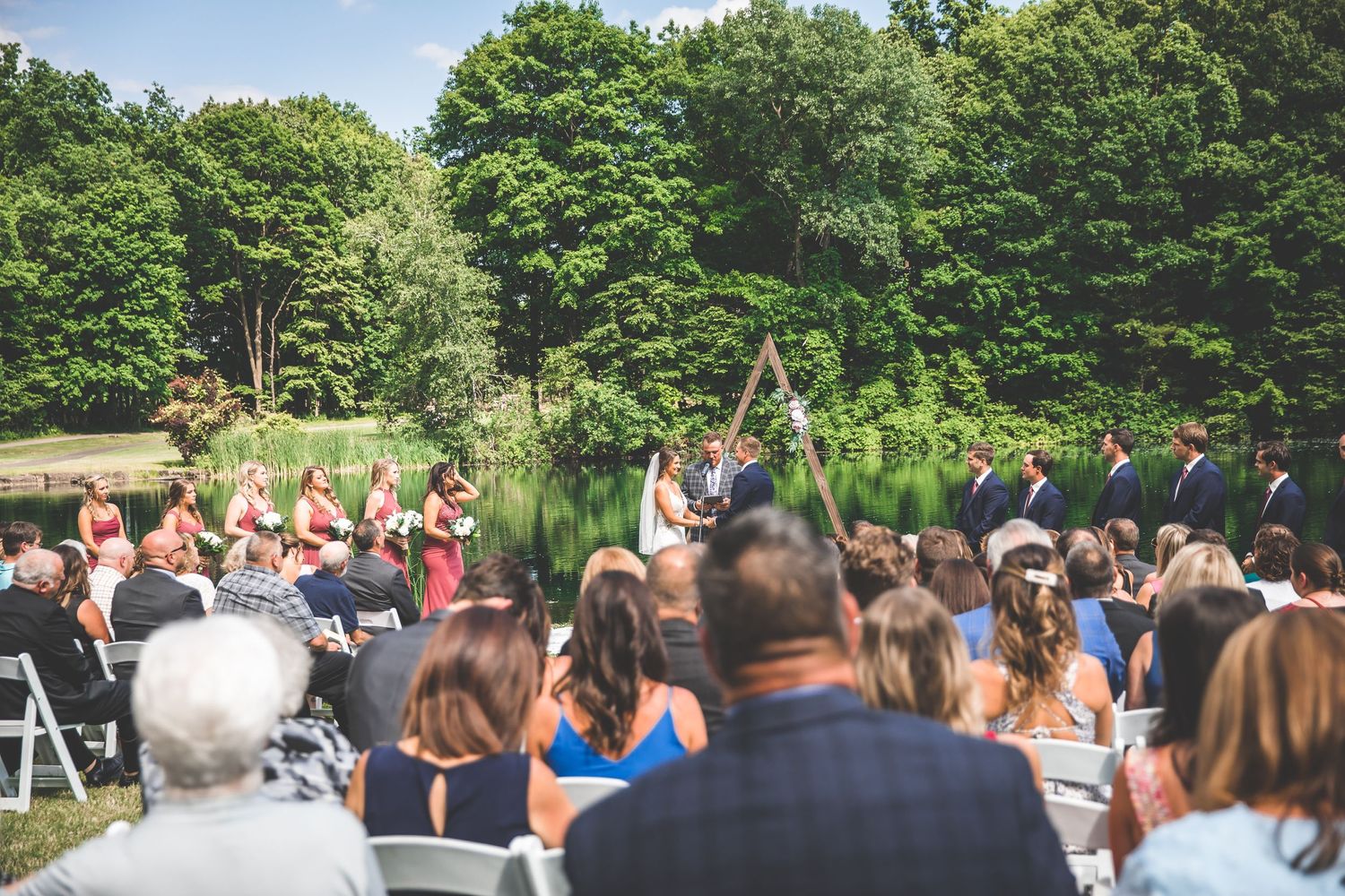 Panoramic view of wedding guests seated in white chairs during outdoor ceremony.
