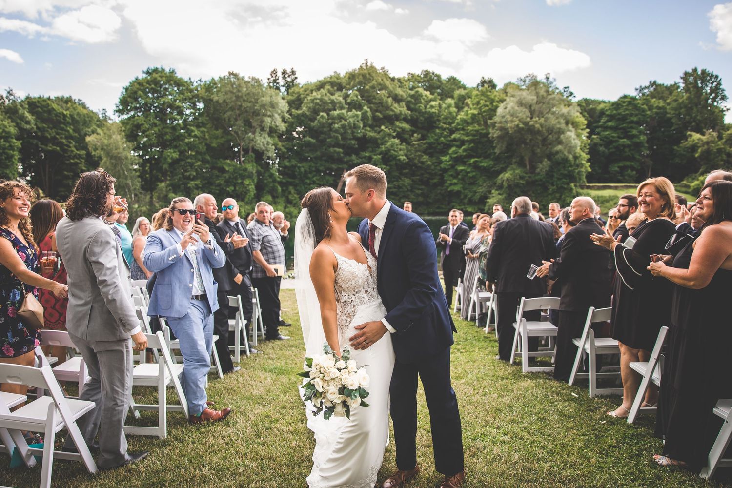 Wedding guests seated on white chairs in grassy field during outdoor ceremony.