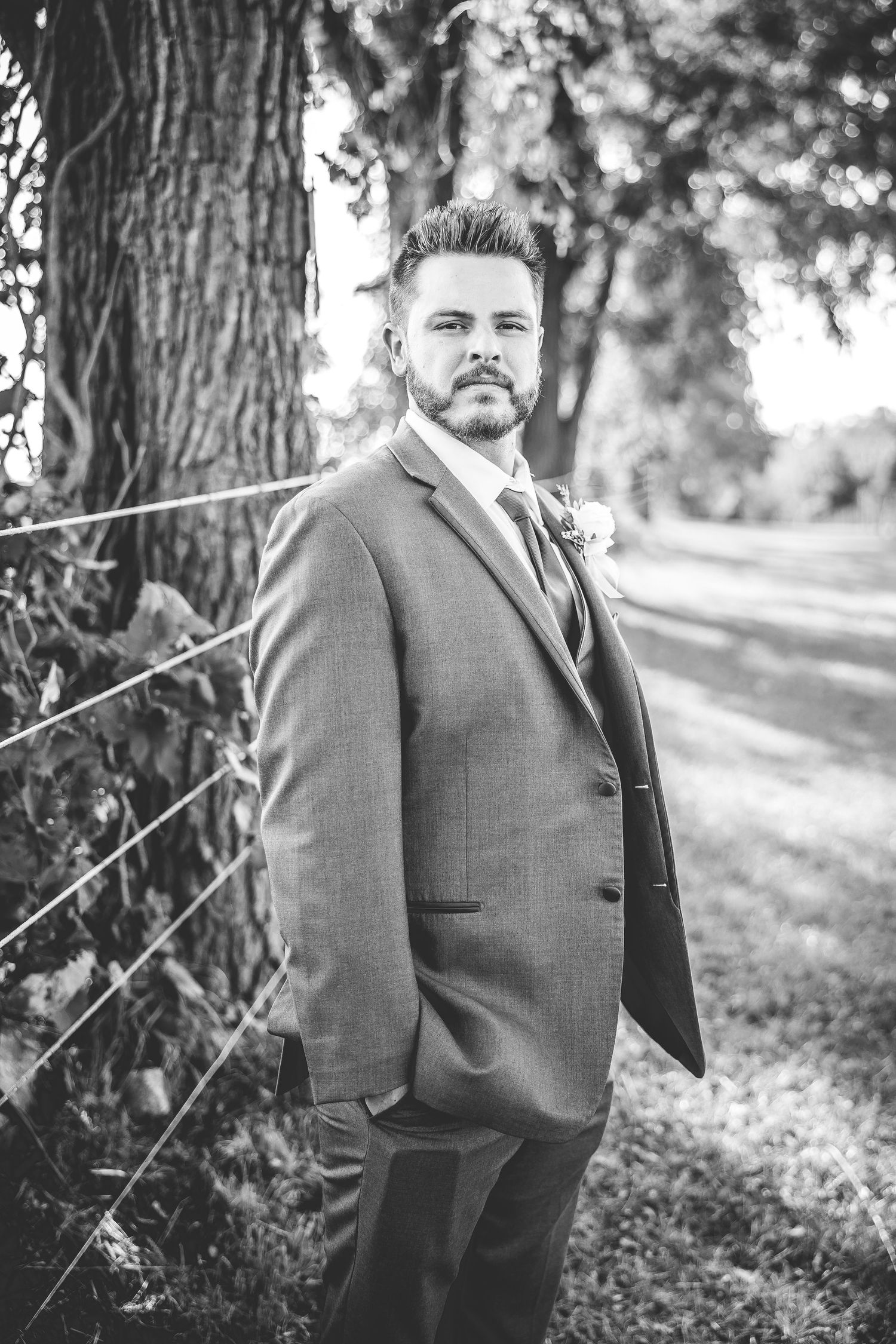 Black and white portrait of groom in formal attire standing beside large tree.