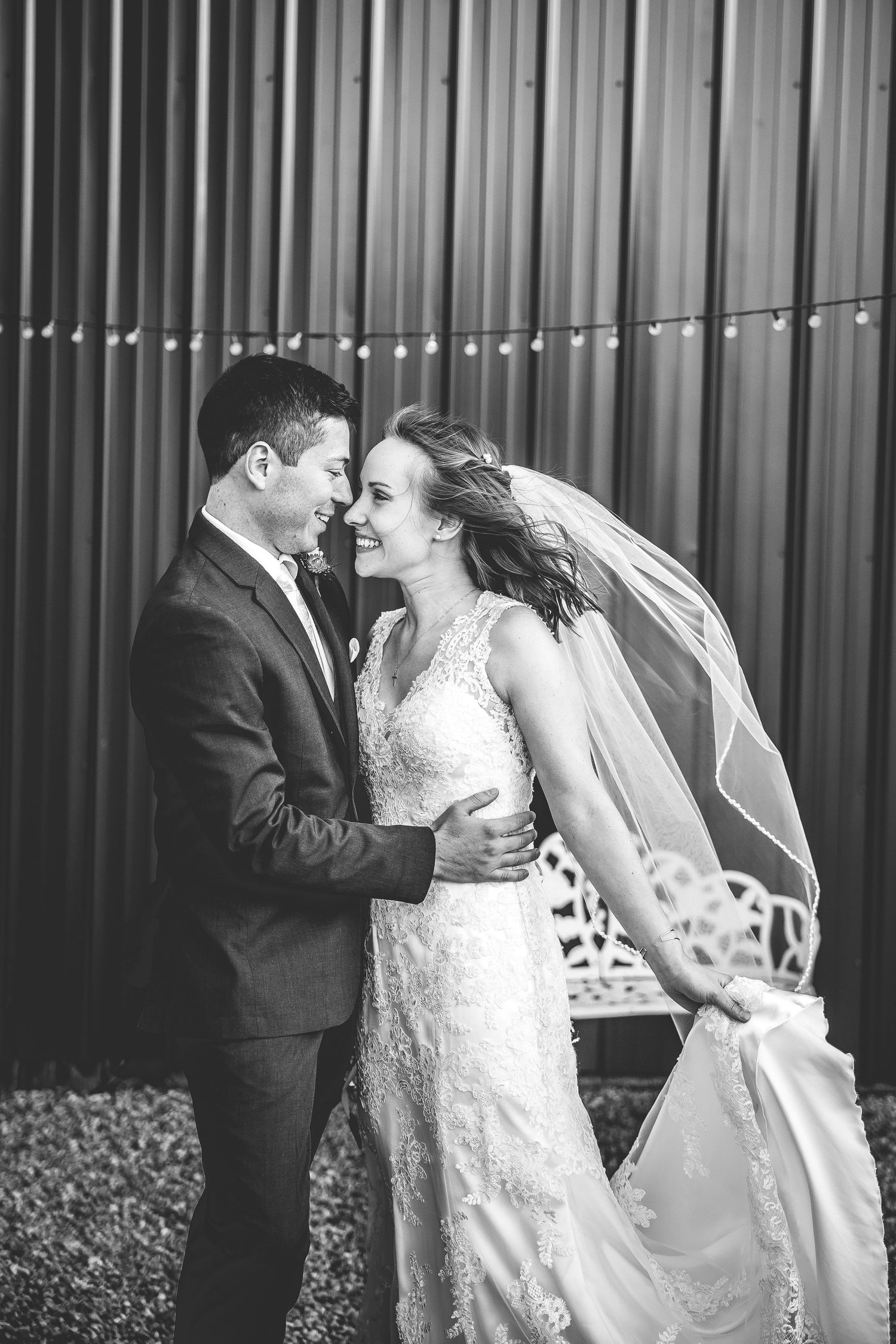 Romantic black and white portrait of couple embracing against industrial backdrop with string lights.