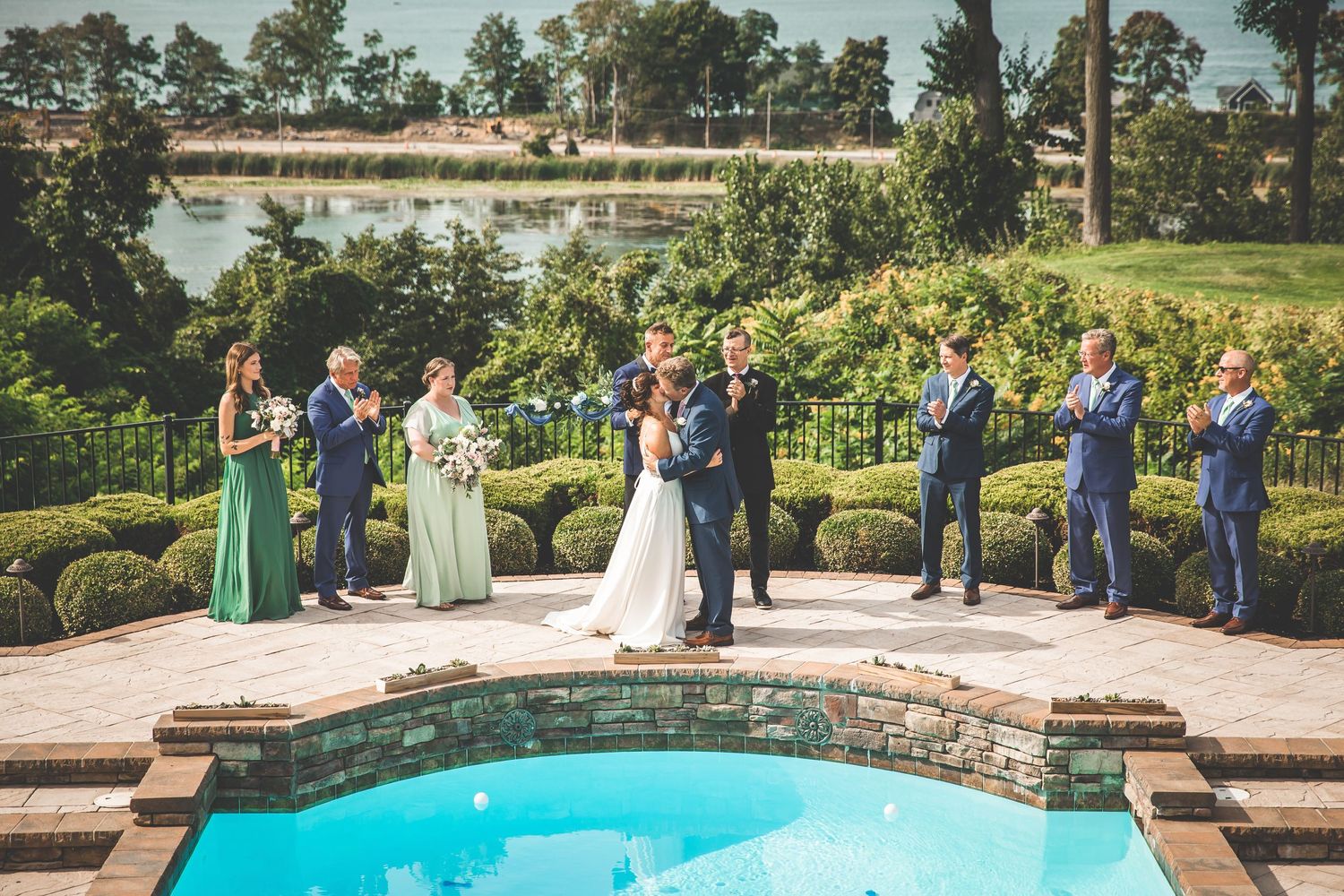 Outdoor wedding ceremony setup with stone wall and swimming pool surrounded by lush landscaping.