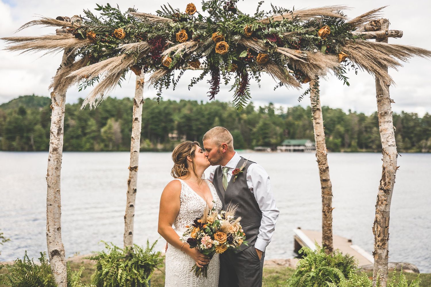 Wedding ceremony site features natural birch poles and floral arrangements by the water.