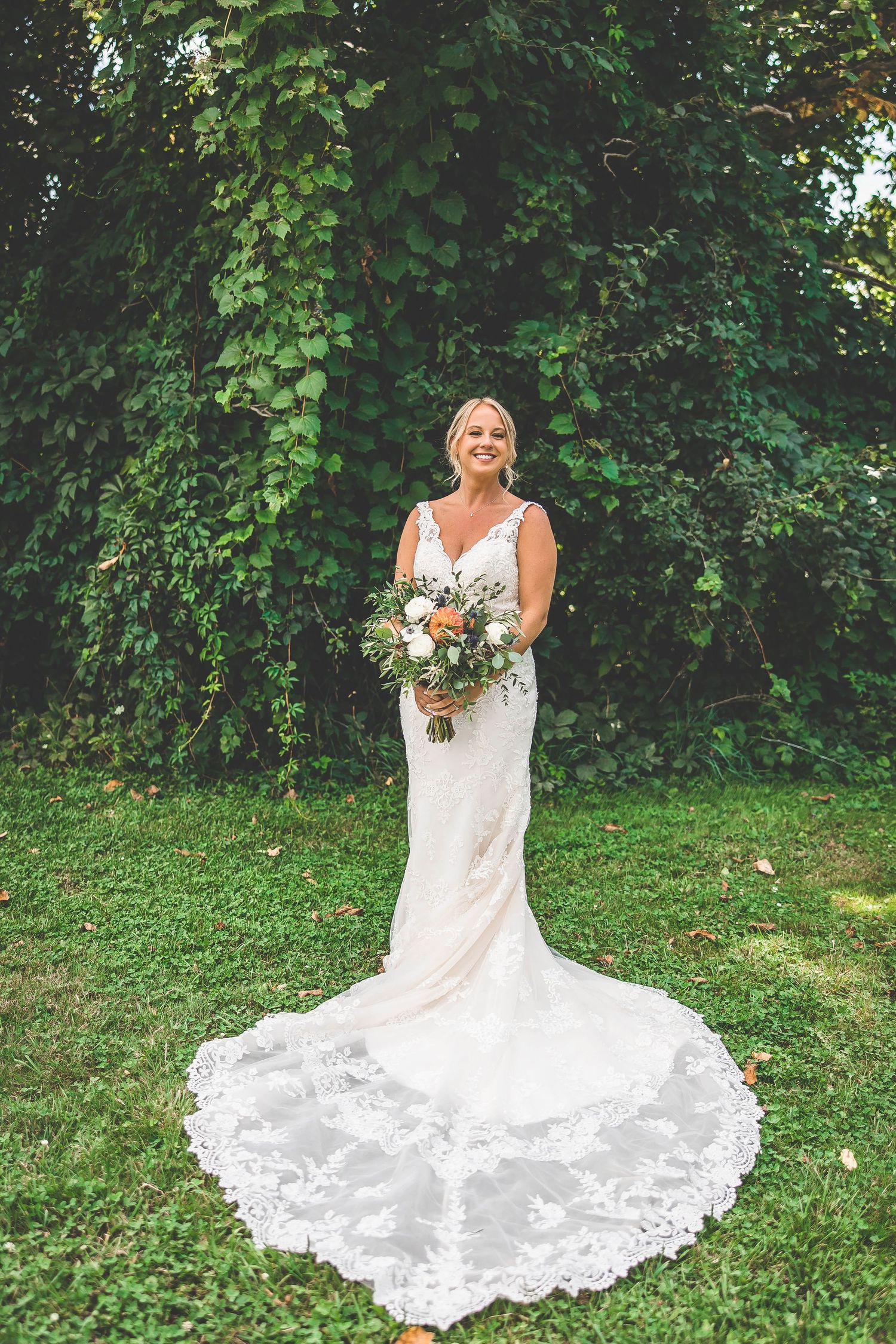 Bride in lace wedding dress sits gracefully on green grass with flowing white train spread around her.