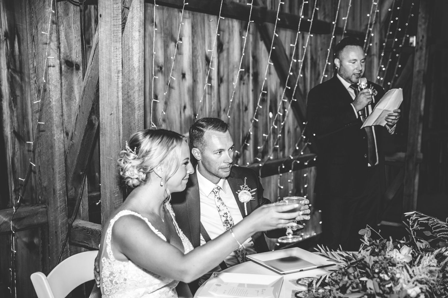 Black and white capture of wedding guests celebrating at tables in a barn reception.