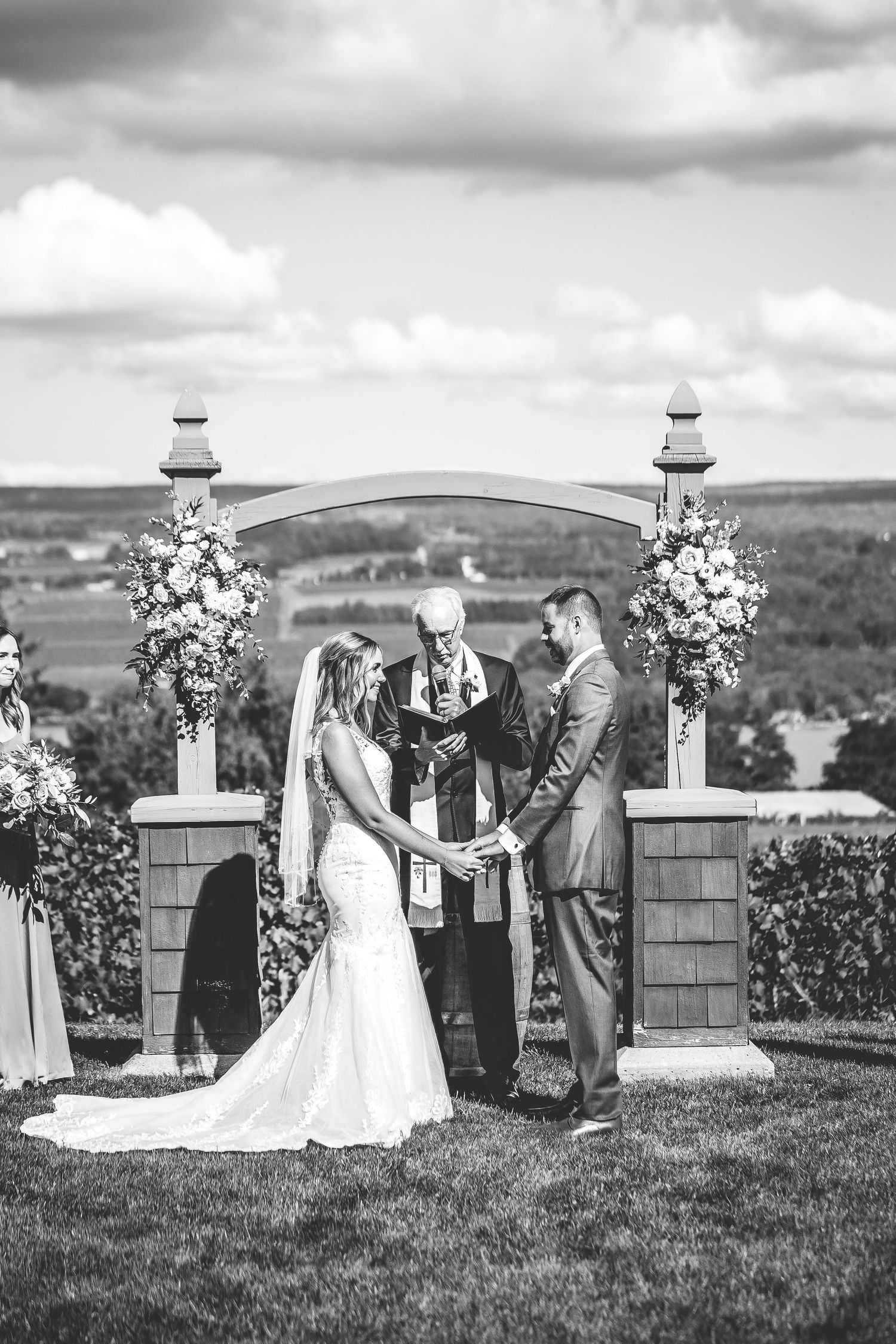 A black and white wedding ceremony under an arch with floral arrangements against a scenic landscape.