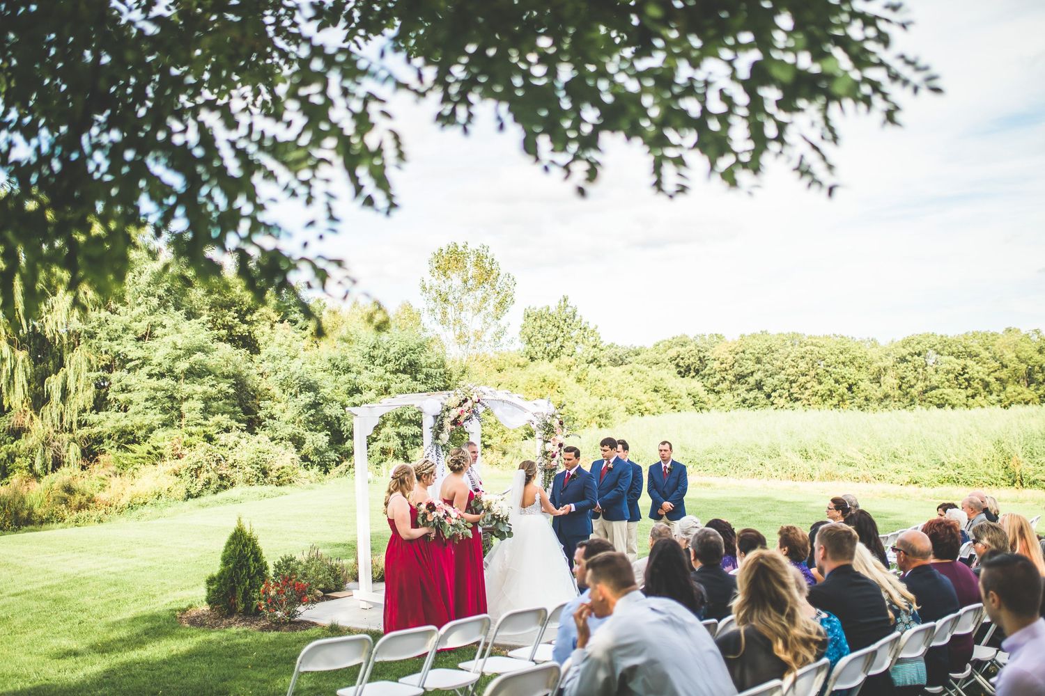 Outdoor wedding ceremony setup with white chairs and guests on a green hillside.