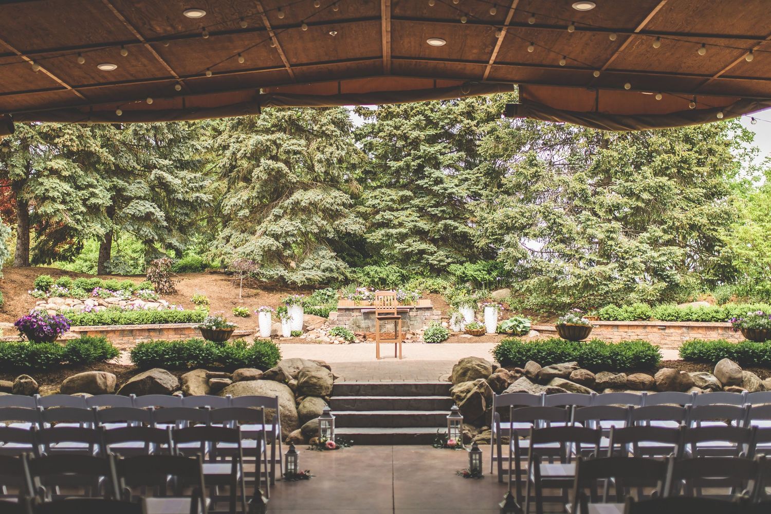 An outdoor wedding ceremony setup with rows of white chairs facing a stage surrounded by lush greenery.