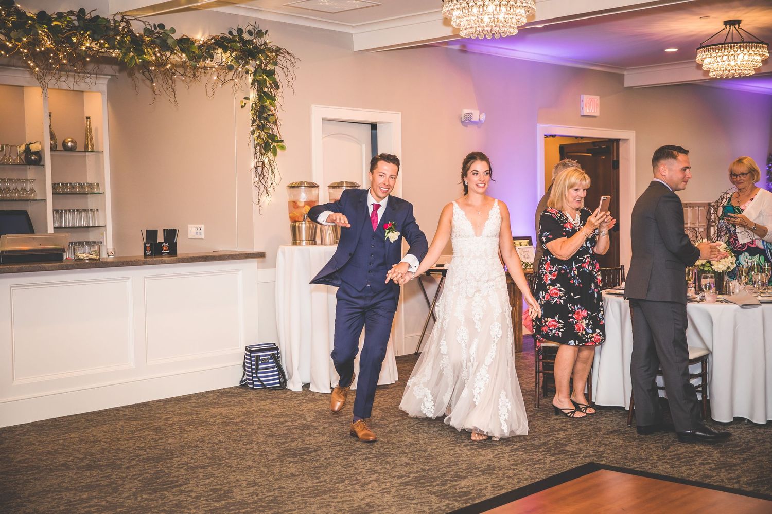 Wedding reception celebration with guests dancing in an elegant venue decorated with chandeliers and purple lighting.