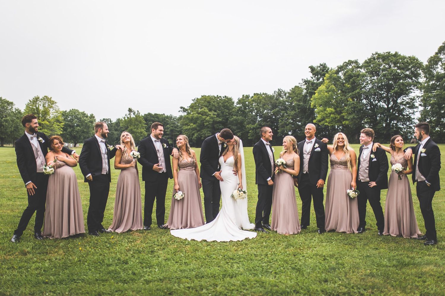 Wedding party in blush pink dresses and black tuxedos poses on grassy hillside.