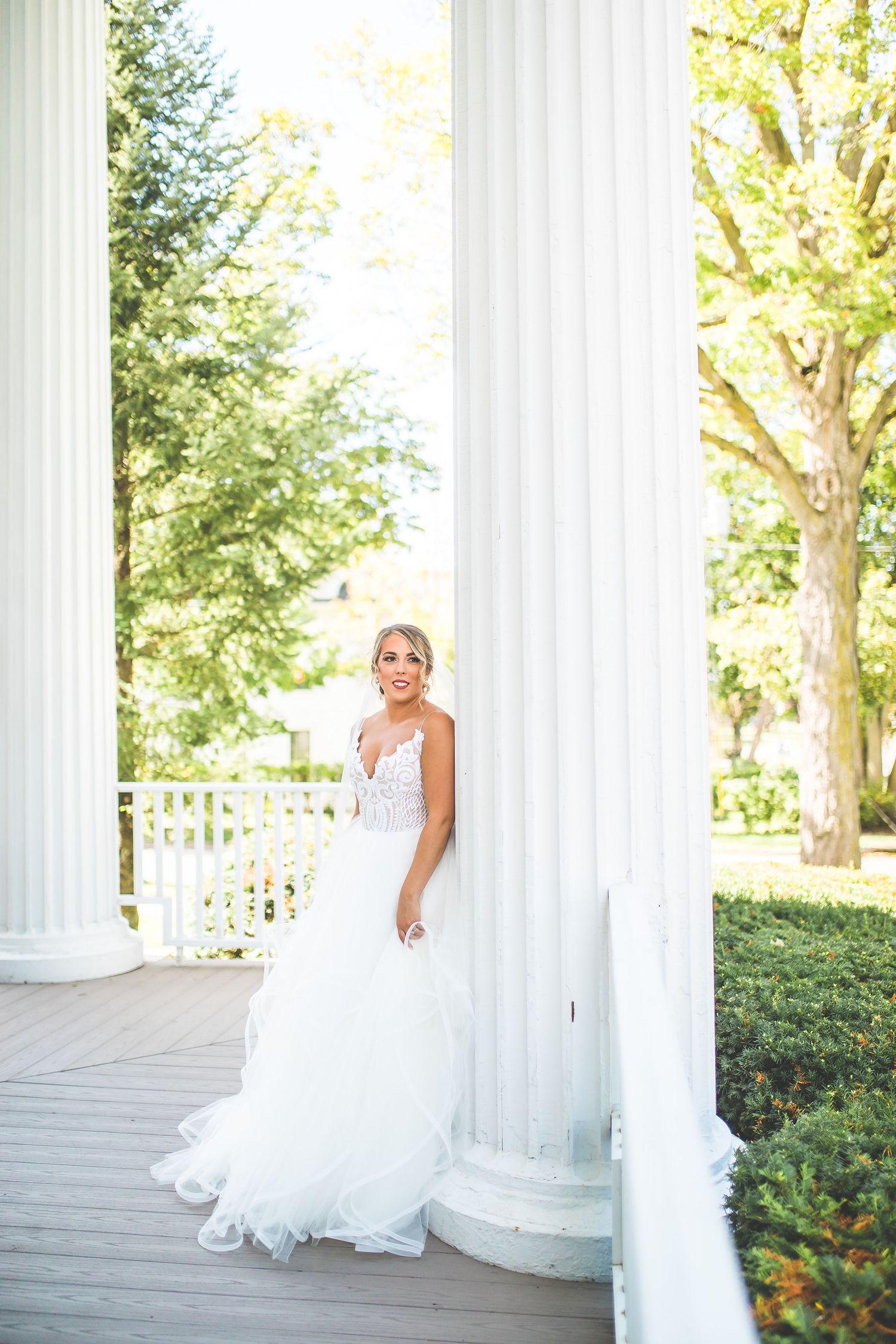 Bride in flowing white gown poses between classic white columns on a sunny day.