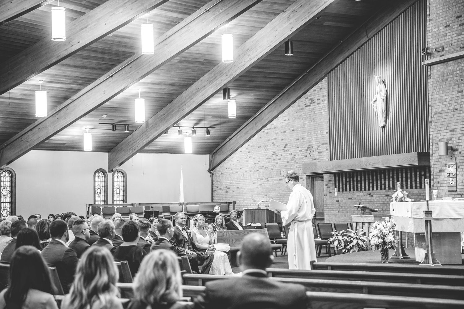 Modern church interior with exposed ceiling beams and pendant lighting during a wedding ceremony.