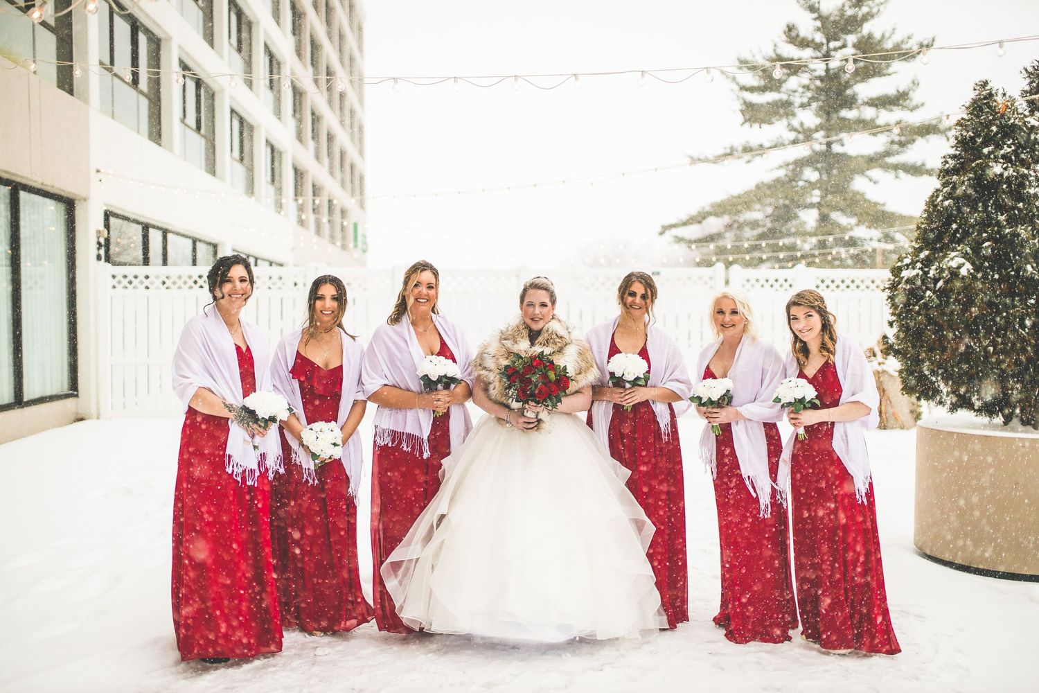 Wedding party in red dresses and navy suits pose together in falling snow.