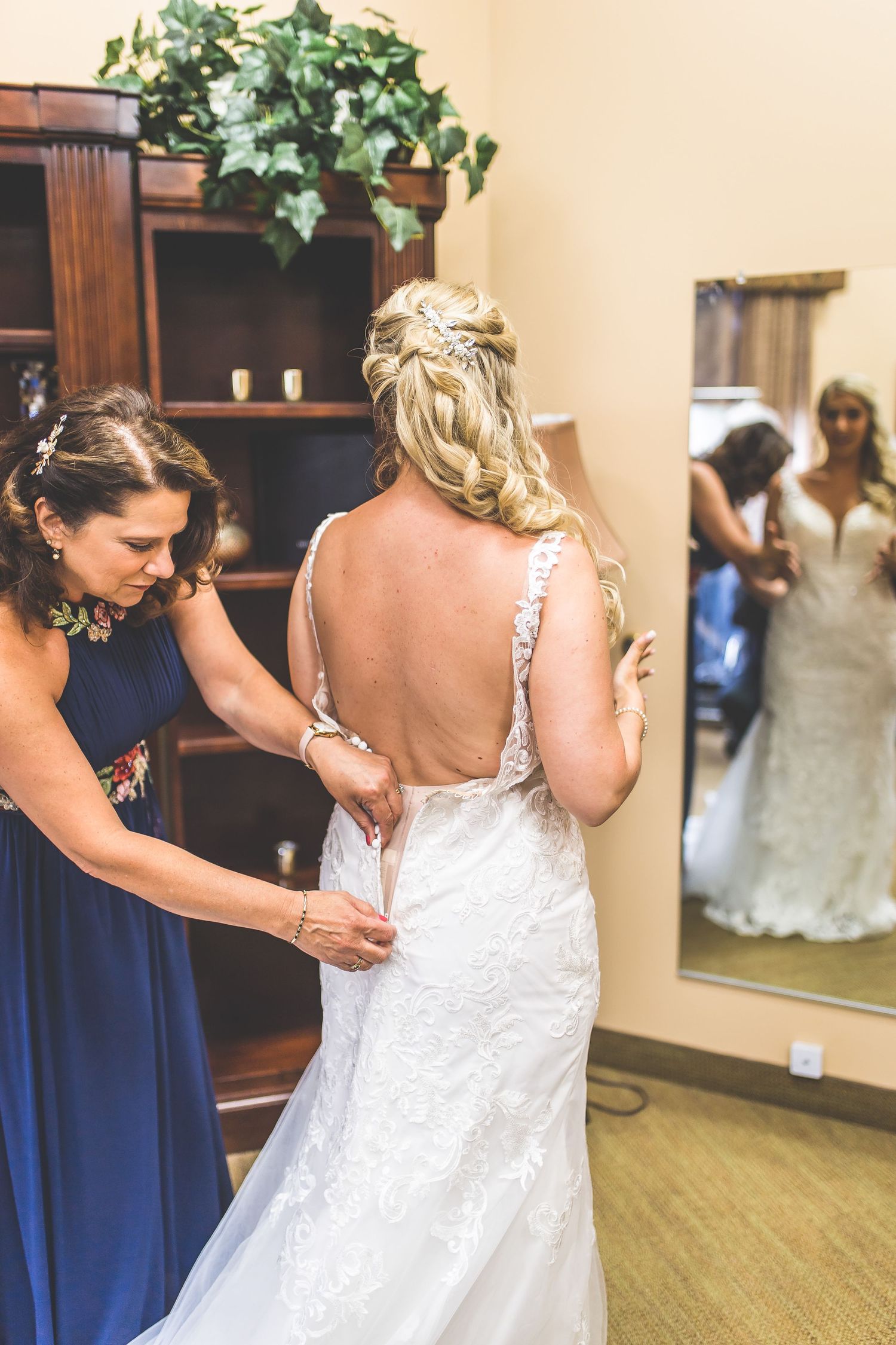 A bridesmaid in navy blue helps adjust the back of a white lace wedding dress during pre-ceremony preparations.