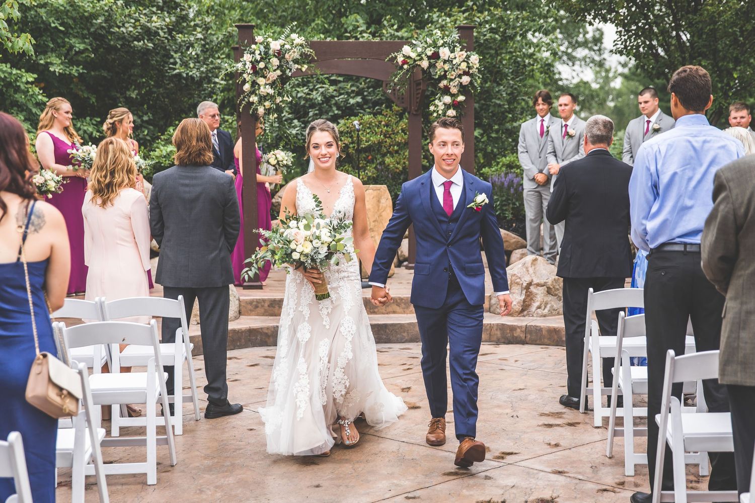 Wedding guests seated in white chairs at outdoor ceremony venue with natural landscaping.