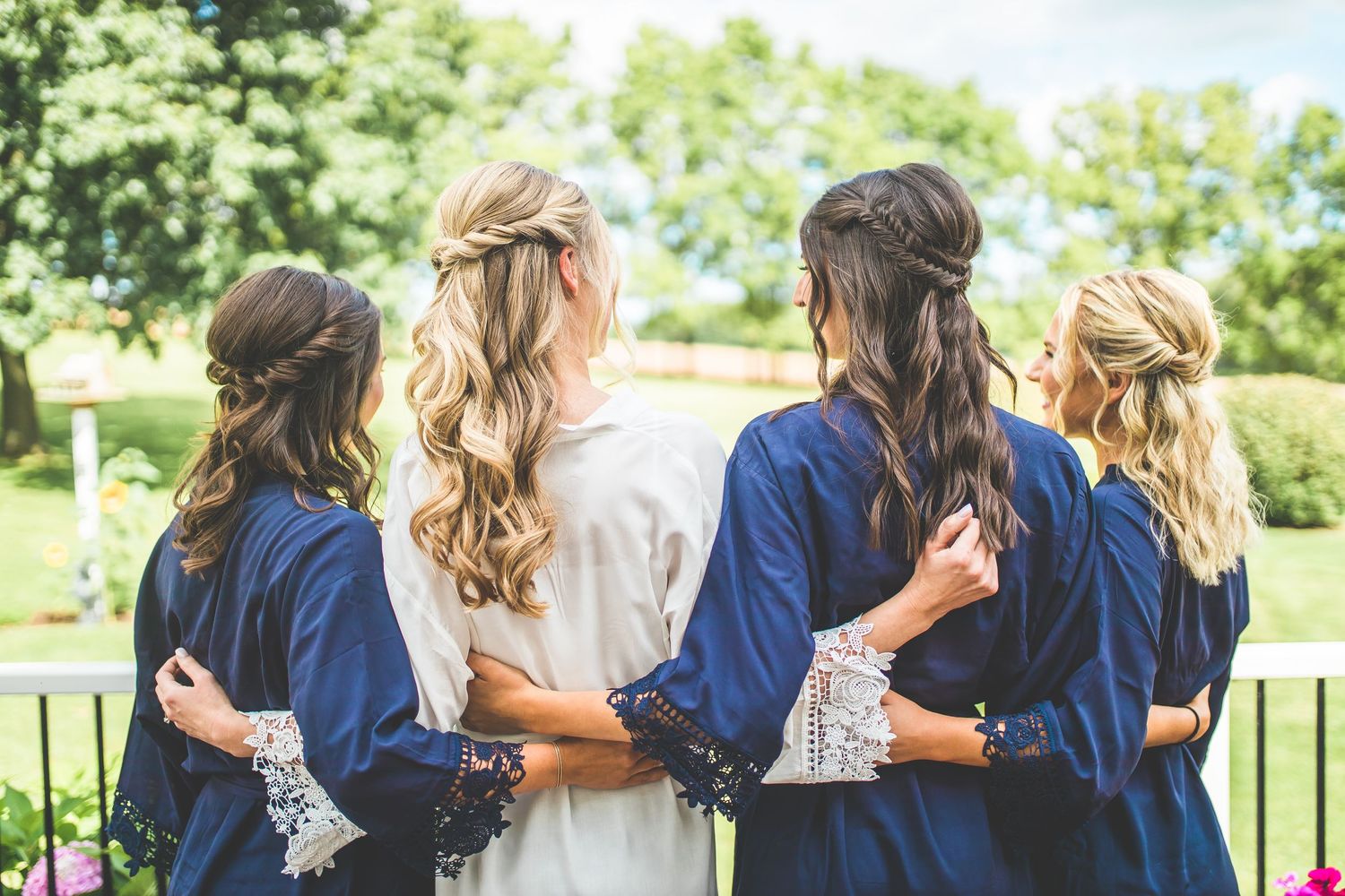 Bridesmaids in navy robes stand together with their backs to the camera in a garden setting.
